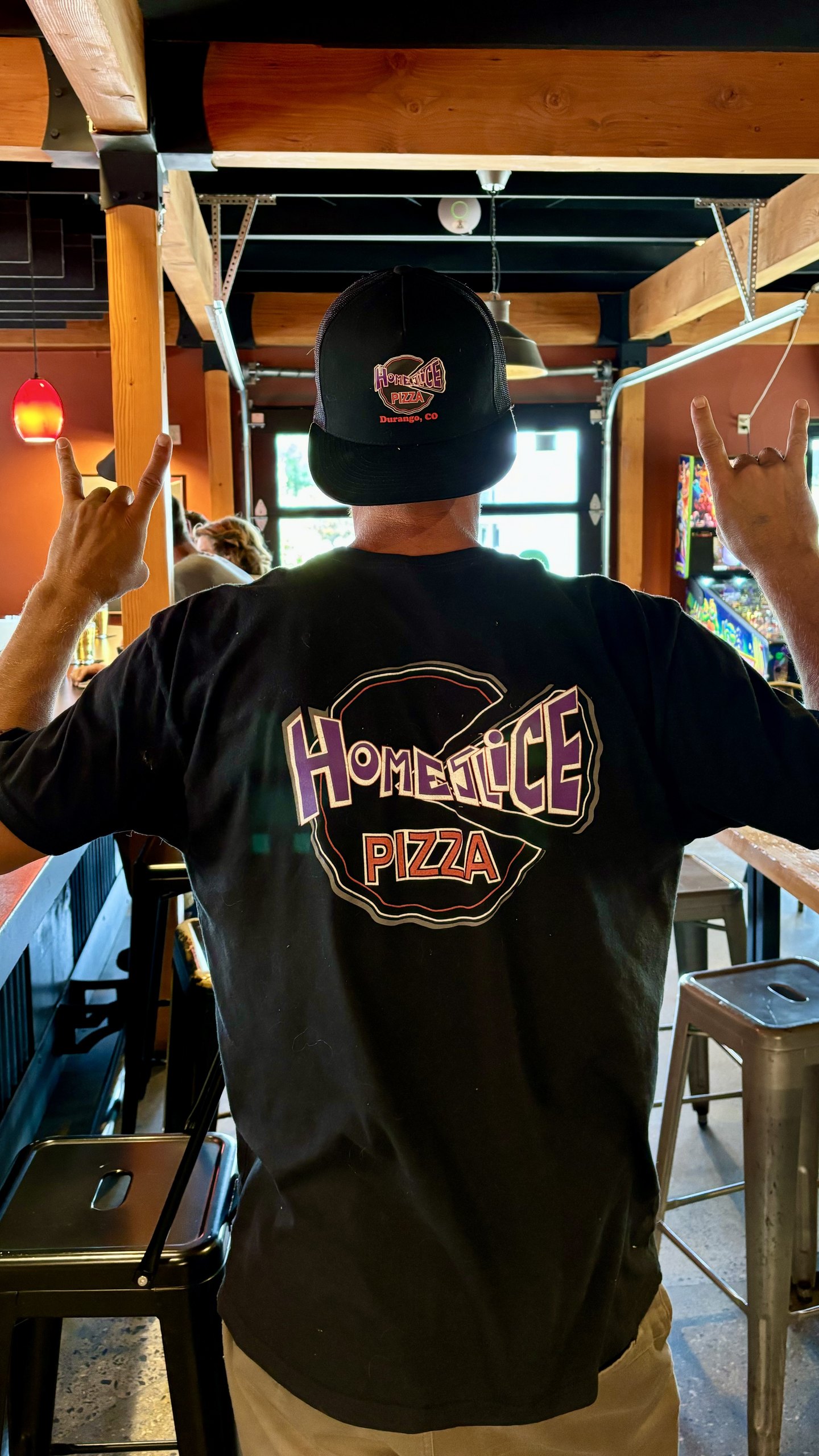 A HomeSlice staffer in a HomeSlice tee throwing rock-on hands, back to camera