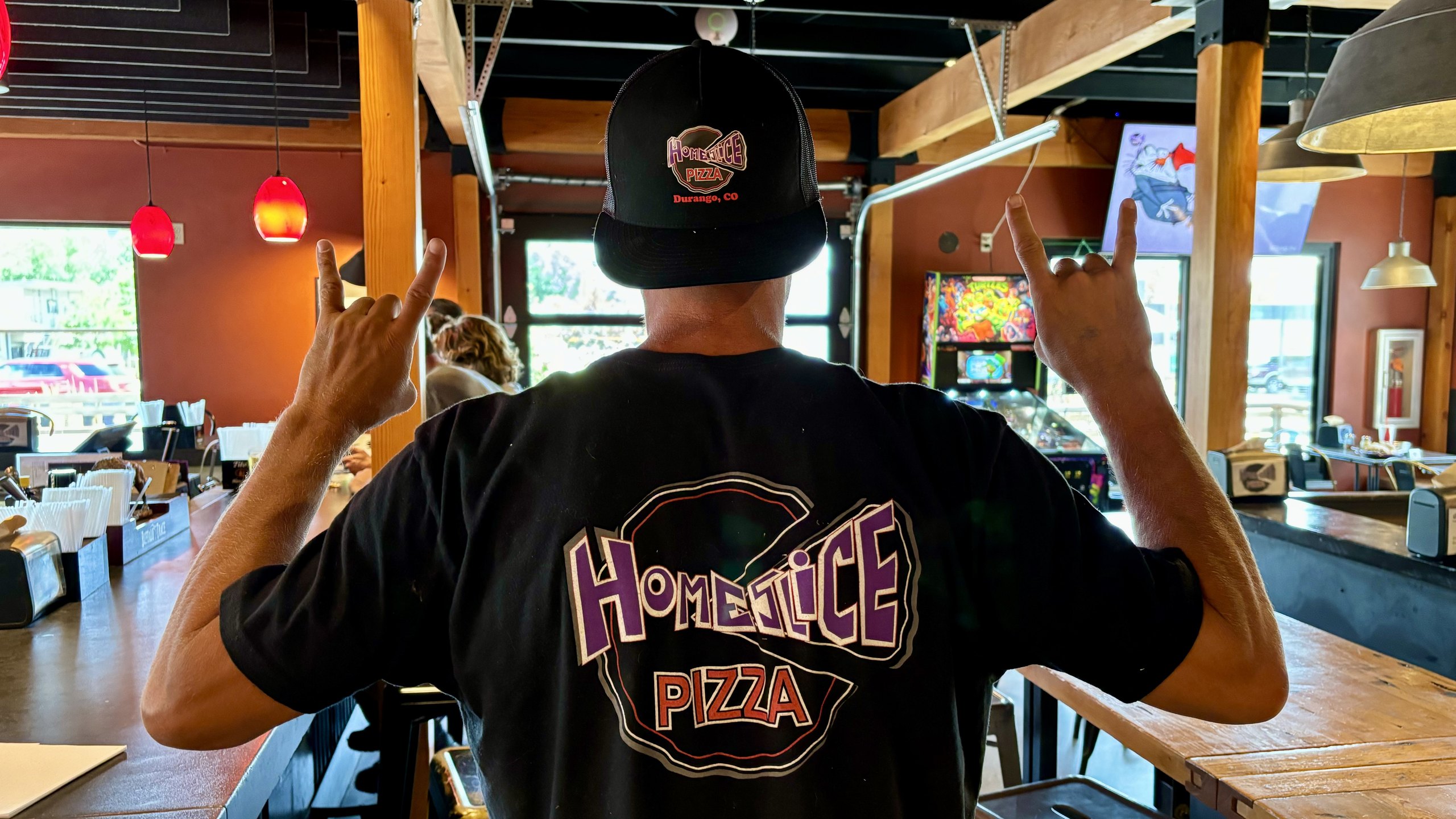 A HomeSlice staffer in a HomeSlice tee throwing rock-on hands, back to camera